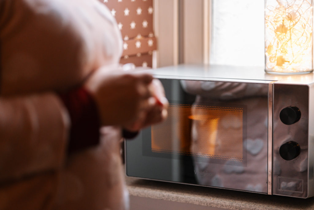 Nieostry pierwszy kadr to. kobieta skłądająca ręce razem a w tle mikrofalówka podgrzewająca posiłek