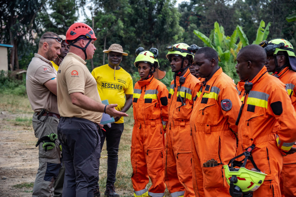 In the photo, PCPM instructors are training a Kenyan USAR team. Male and female firefighters are standing in a line facing the PCPM instructors and listening attentively to the training