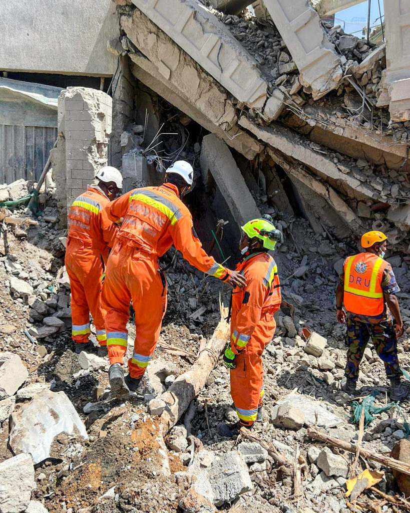 Four firefighters are amid rubbles of a collapsed building in Nairobi, wearing orange suits and discussing further actions